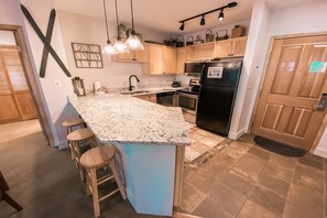 A kitchen with a granite countertop, wooden stools, a black refrigerator, and wooden cabinets.