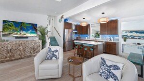 A modern living room with a white sofa, a coffee table, and a TV displaying a beach scene. A kitchen with wooden cabinets and a bar area is visible in the background. - Bluewater Resort is 2 Properties Sleeping up to 14 (Fort Myers Beach)