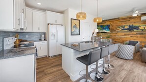 A modern kitchen with white cabinets, a central island with bar stools, wooden accents on the walls, and pendant lighting. - Bluewater Resort is 2 Properties Sleeping up to 14 (Fort Myers Beach)