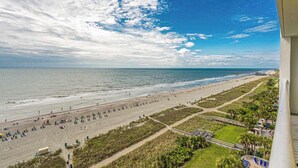 A beach with a long stretch of sand, a pier, and a path leading to a building with a balcony.