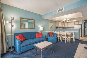 A room with a blue sofa, a small table, a dining area with chairs, a kitchen with white cabinets, and a mirror on the wall. - Ocean Front Views! Camelot Resort 1011 (Myrtle Beach)