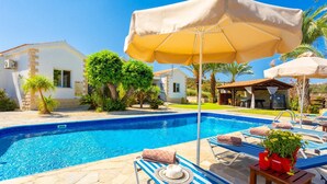 A swimming pool with lounge chairs and umbrellas, surrounded by greenery and a stone building. - Villa Amorosa in Neo Chorio (Neo Chorio)