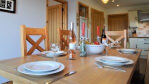 Een eettafel gedekt met borden, wijnglazen en flessen, met houten stoelen en een keuken op de achtergrond.