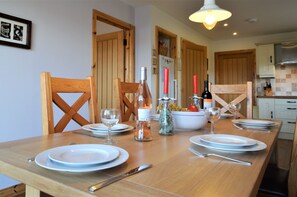 A dining table set with plates, wine glasses, and bottles, with wooden chairs and a kitchen in the background. - High Beach Cottage, coastal, picturesque, sea views (Kames, near Tighnabruaich)