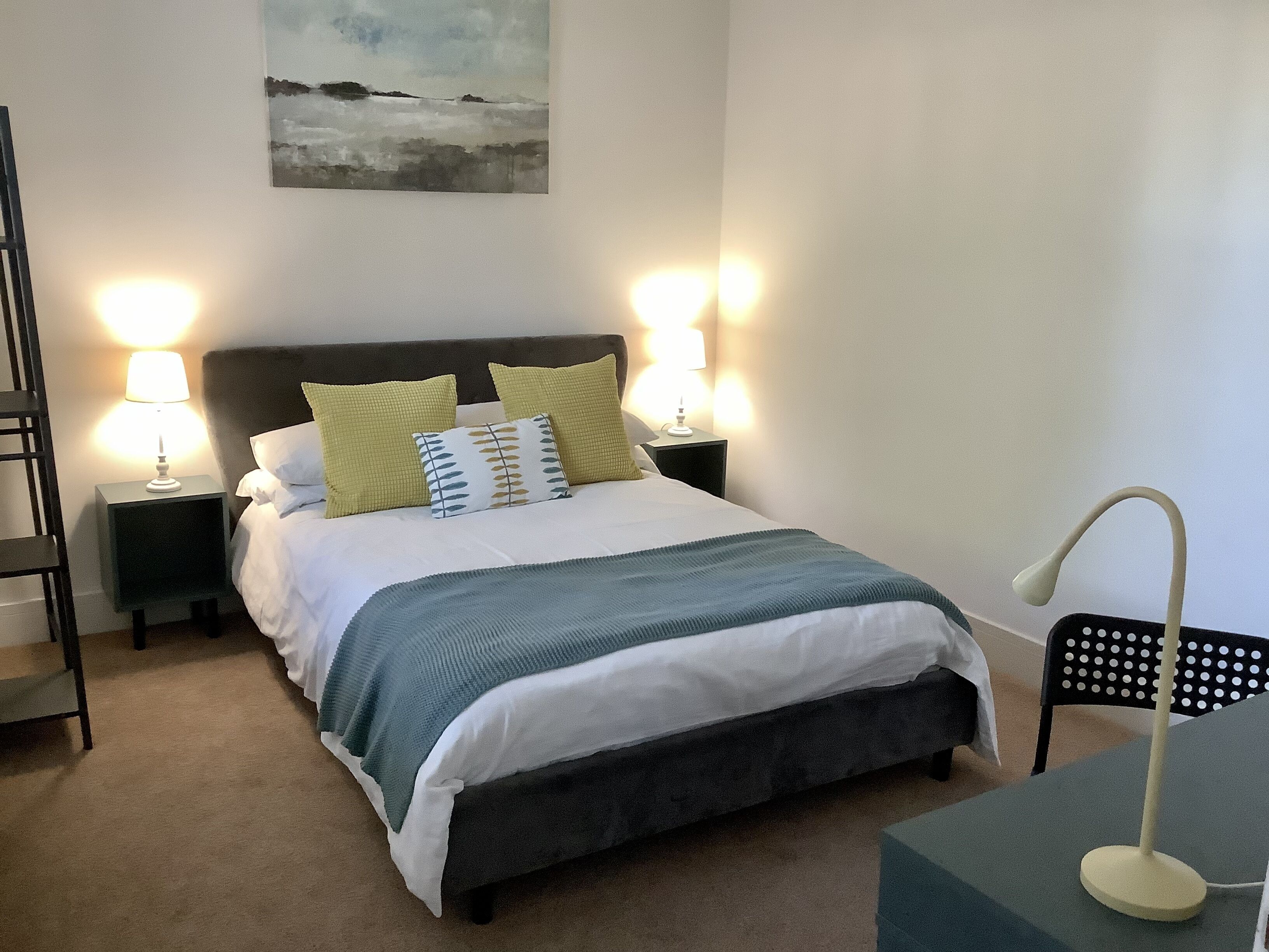Un lit bien fait, avec une tête de lit sombre, deux tables de chevet avec des lampes, une chaise et un tableau accroché au mur.