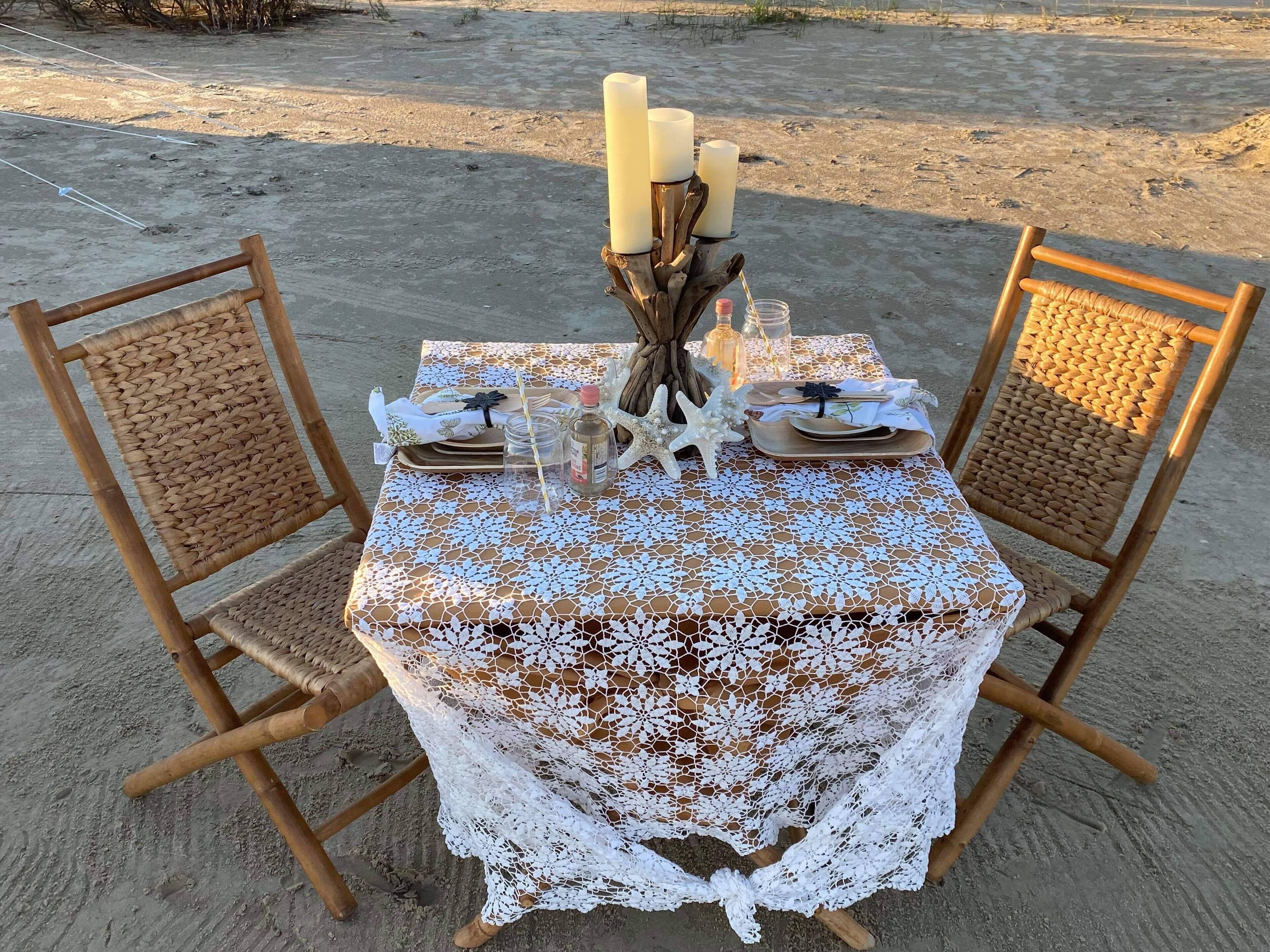 A table set for two with a white lace tablecloth, wooden chairs, and candles.