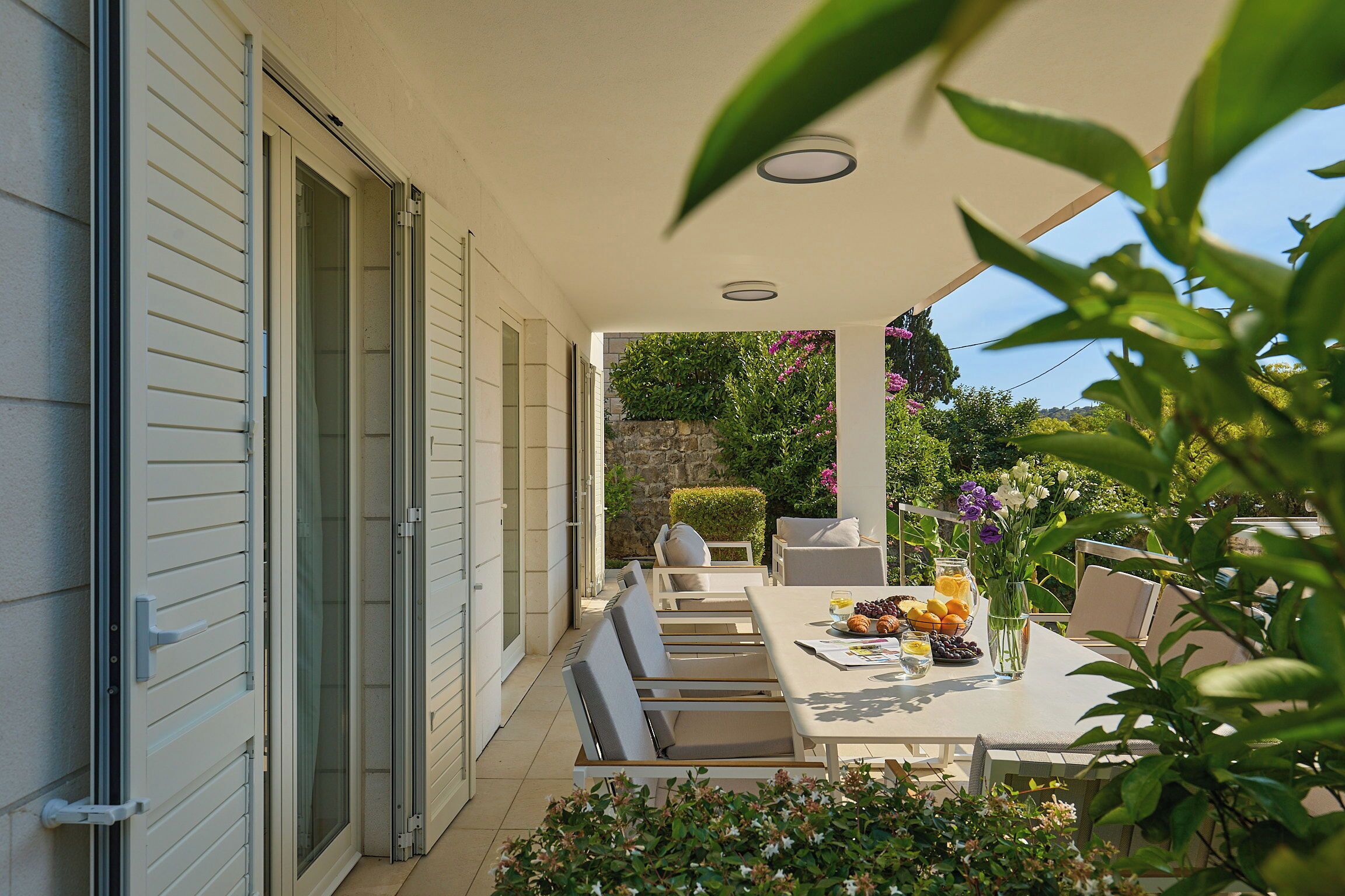 An outdoor dining area with a table set for two, surrounded by greenery and a stone wall in the background.
