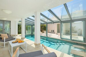 A modern indoor pool area with glass walls, a lounge area with seating, and a table with fruits and drinks. - Hedera Estate, Villa Hedera III (Zaton)