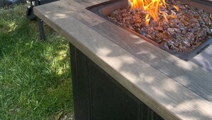 Un braciere con fiamme, circondato da una panca in legno e un albero.