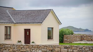 A single-story house with a stone wall in front, a dark brown door, and a view of the sea and hills in the background.