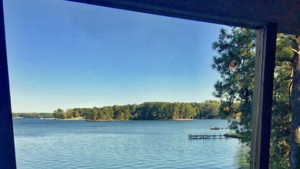 Eine Holzterrasse mit einem Stuhl, mit Blick auf einen stillen See mit einem Steg und Bäumen im Hintergrund.