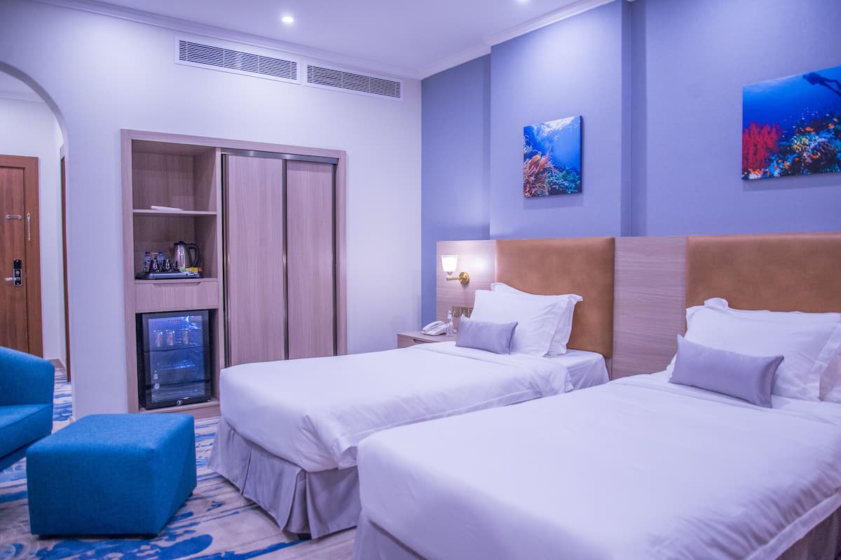 A hotel room with two beds, a minibar, a desk with a lamp, and artwork on the walls.