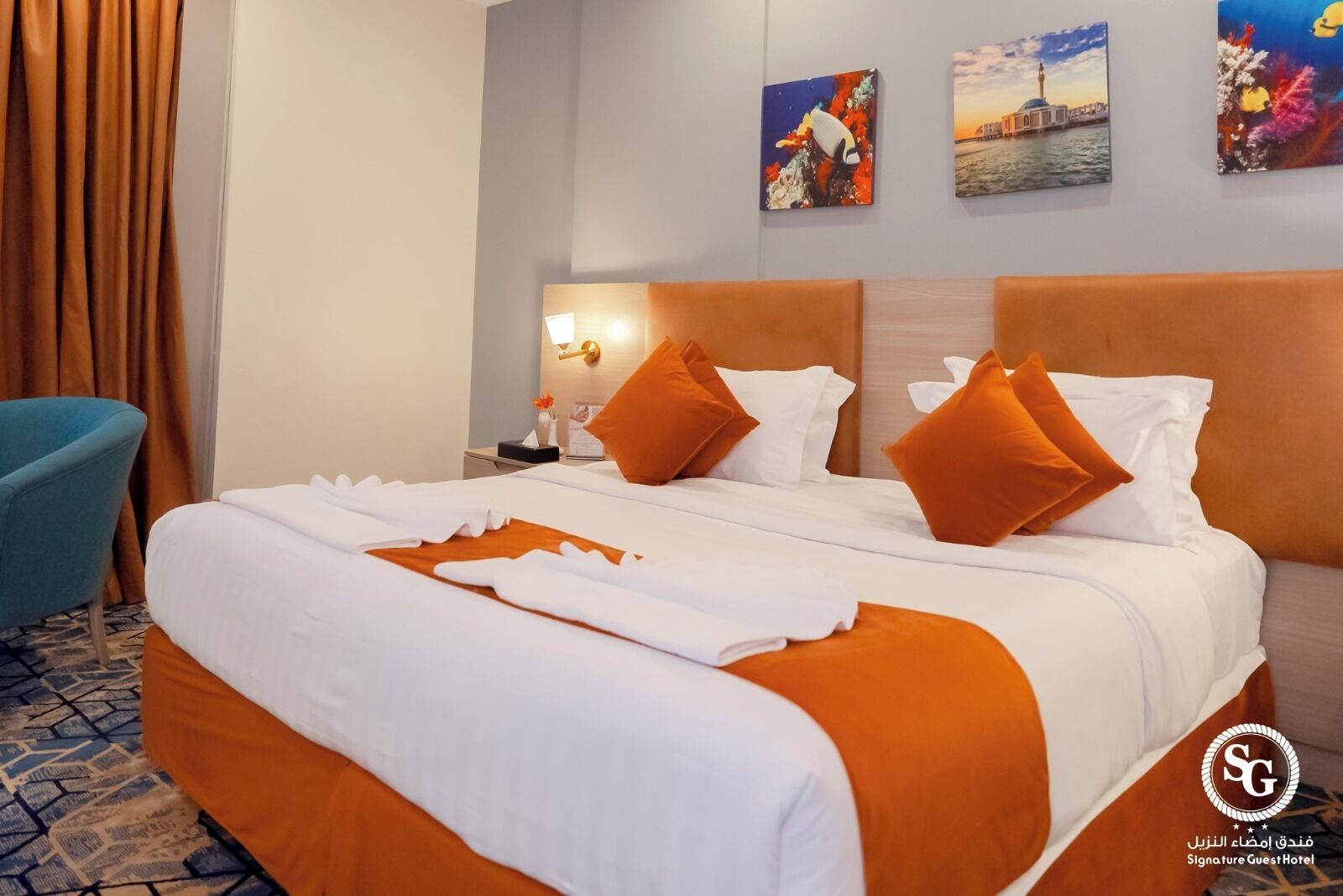 Una habitación de hotel con una cama grande, ropa de cama naranja, una mesita de noche con lámpara y una obra de arte en la pared.