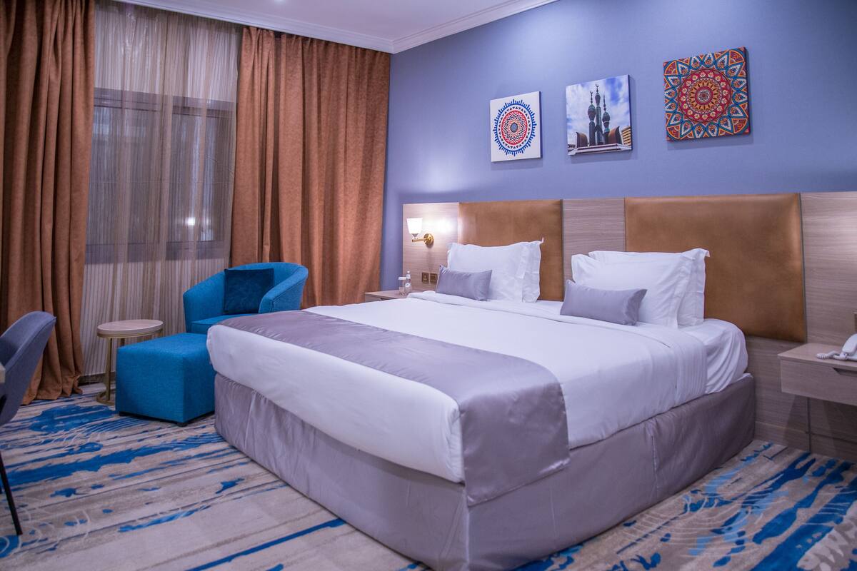 A hotel room with a large bed, a blue armchair, a small round table, and a nightstand with a lamp.