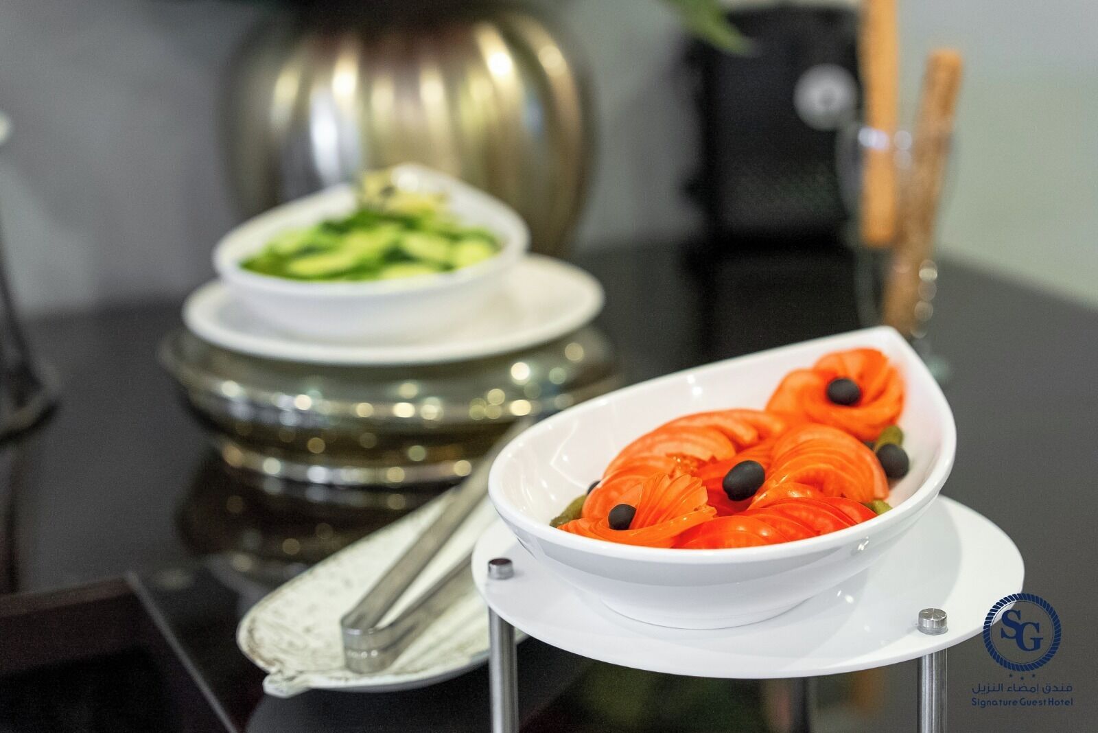 A table with three bowls of food, a pair of tongs, and a decorative vase.