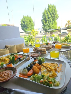 A breakfast buffet with various dishes and drinks on a table, set against a backdrop of greenery and a building. - فندق إمضاء النزيل الفندقية (Jeddah)