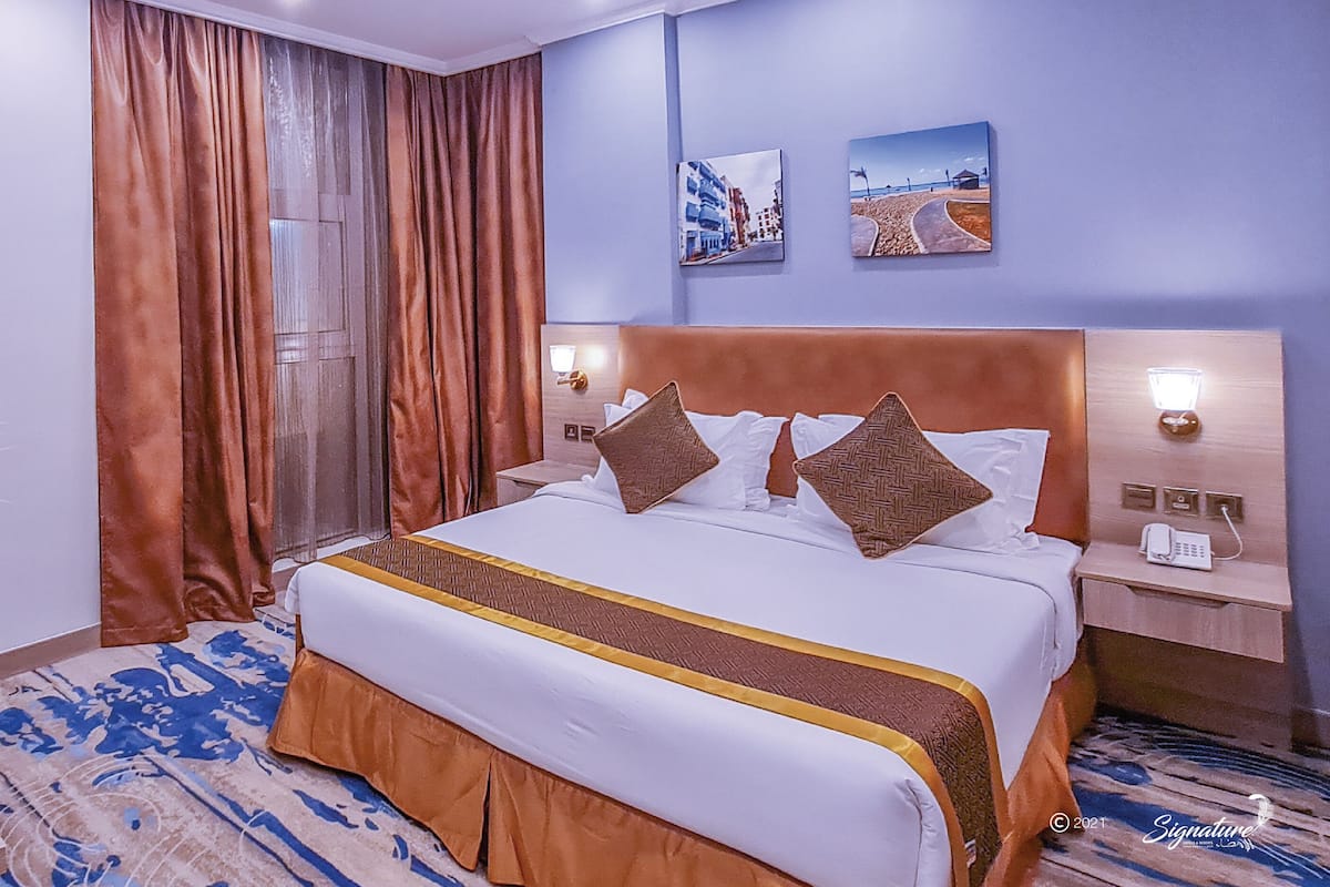 A hotel room with a large bed, a desk with a lamp, a chair, and a view of the city through the window.