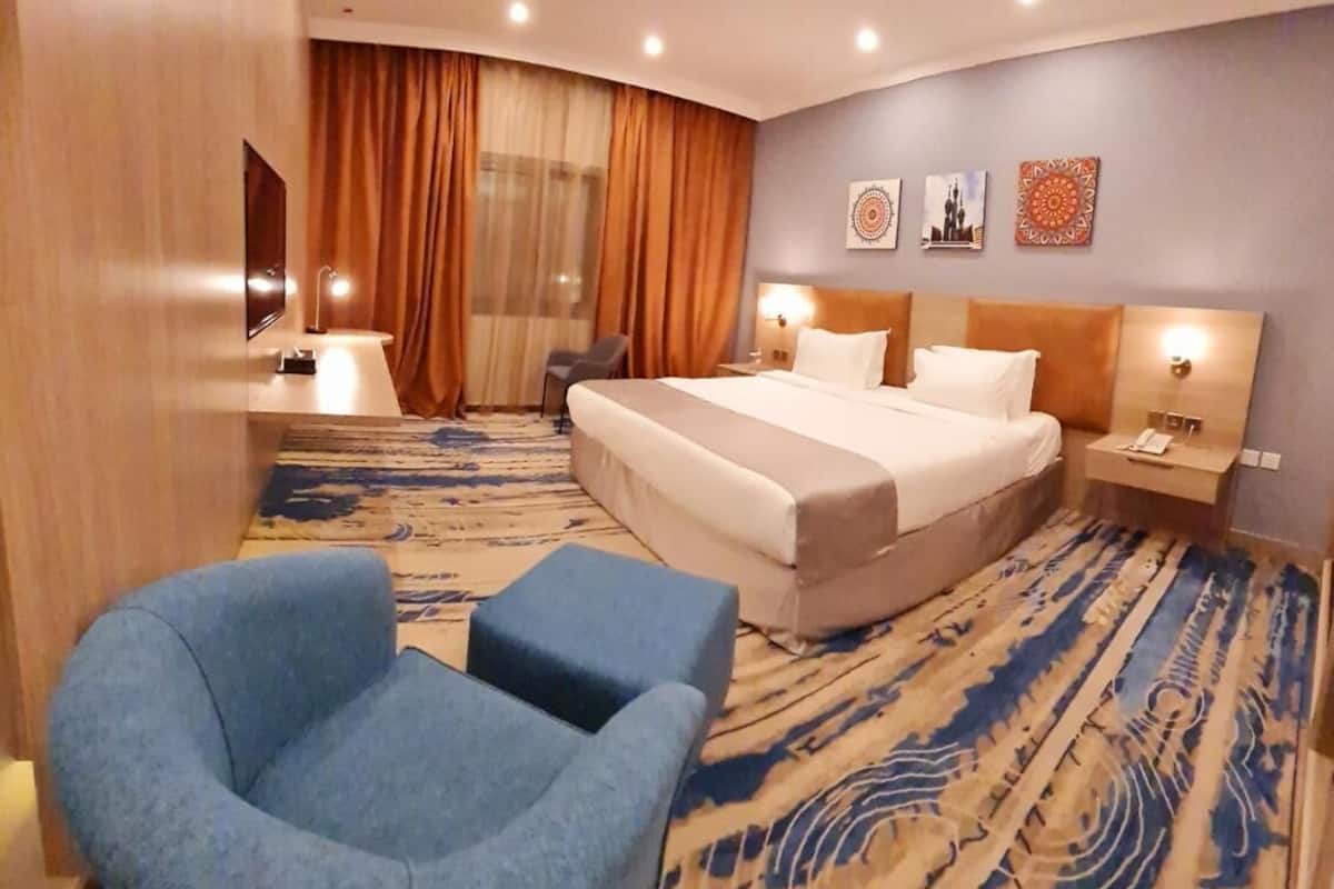 A hotel room with a large bed, a blue armchair, a desk with a lamp, and a view of the cityscape through the window.