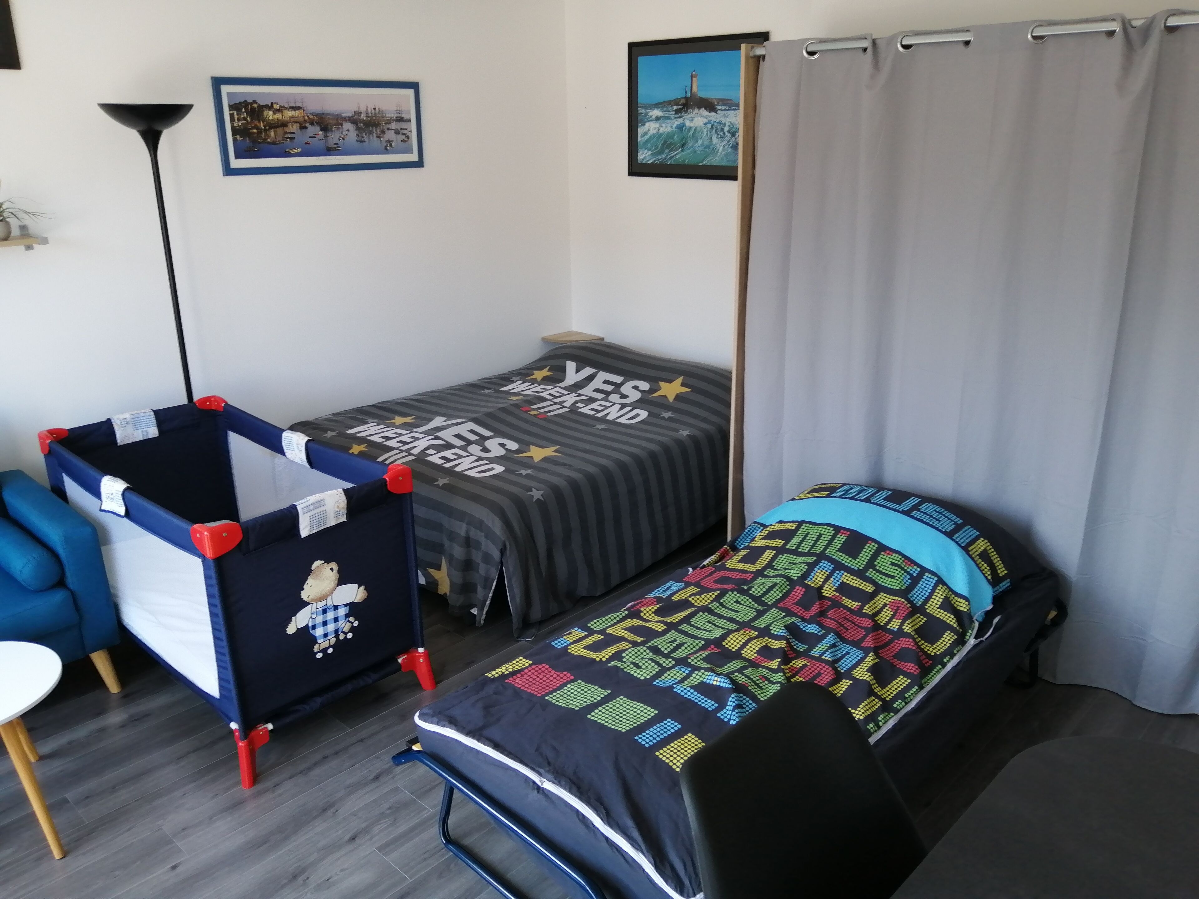 Quarto pequeno e bem arrumado com duas camas de solteiro, uma cadeira azul, uma mesinha e um cercadinho.