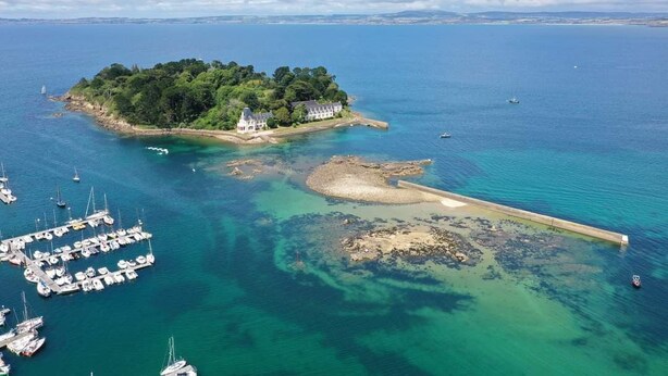 Uma ilha com um edifício branco, rodeada por vegetação exuberante e águas azul-claras.