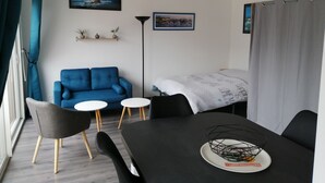 Um espaço de estar moderno e compacto, com um sofá azul, uma pequena mesa de jantar e uma cama com um cobertor estampado em branco.