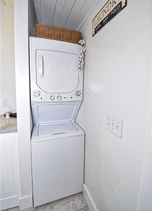 Une petite buanderie avec un lave-linge et un sèche-linge blancs, un panier posé sur le sèche-linge et une pancarte au-dessus indiquant « BUANDERIE - PLIER ».