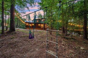 A rustic cabin with a swing set and string lights in a wooded area.