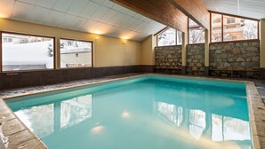 Una piscina con paredes de piedra y vigas de madera, ventanas amplias y vistas a un paisaje nevado.