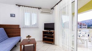 A room with a bed, a TV on a wooden stand, a small table with a vase, and a balcony with chairs and a table. - Holiday apartment with mountain view (Baška)