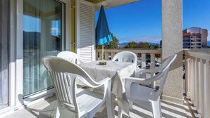 A balcony with a table and chairs, a blue umbrella, and a view of a parking area and buildings. - Holiday apartment with mountain view (Baška)