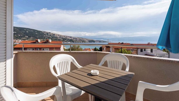 Un balcon avec une table en bois et des chaises blanches, donnant sur une ville côtière aux bâtiments aux toits rouges et offrant une vue sur la mer.