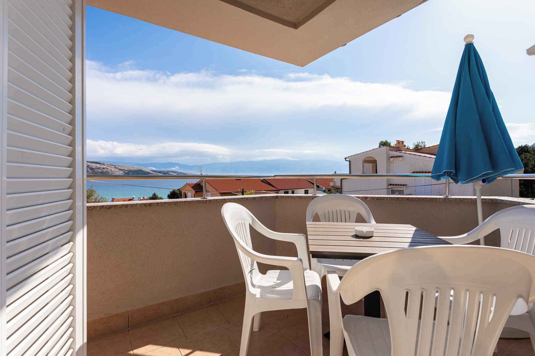Un balcon avec des chaises et une table en plastique blanc, offrant une vue panoramique sur la mer et les montagnes lointaines.
