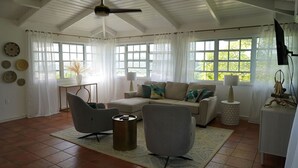 A spacious living room with a high ceiling, a ceiling fan, a beige sofa with patterned cushions, two armchairs, a coffee table, and a large window with curtains. - St. Croix - Villa Views at Hermon Hill! (St Croix)