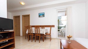 Un coin repas avec une table et des chaises, un téléviseur posé sur un meuble, et une vue sur l’extérieur à travers une porte vitrée.