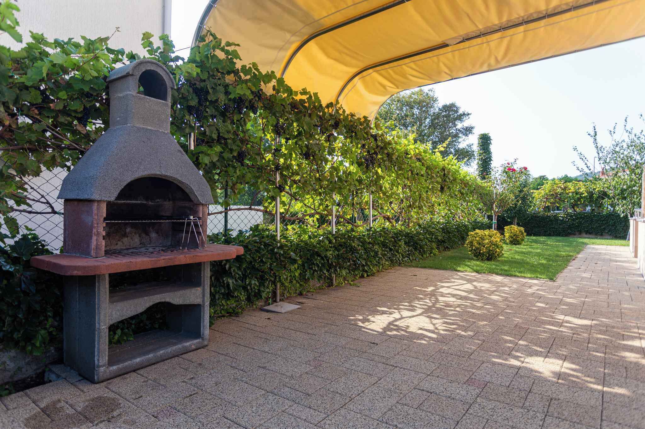 An outdoor brick oven with a chimney, surrounded by a hedge and a yellow canopy.