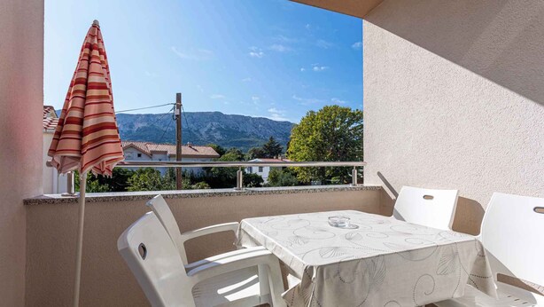 Ein Balkon mit Tisch und Stühlen, einem gestreiften Sonnenschirm und Blick auf Berge und Häuser.