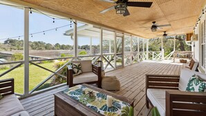 A covered outdoor area with wooden flooring, multiple seating options including sofas and a bench, ceiling fans, and string lights. - NEW Waterfront, private dock, fully updated Johns Island home (Johns Island)