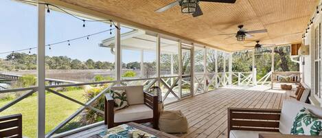 A covered outdoor area with wooden flooring, multiple seating options including sofas and a bench, ceiling fans, and string lights.