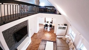 A modern loft apartment with a high ceiling, a fireplace, a dining area, and a staircase leading to an upper level.
