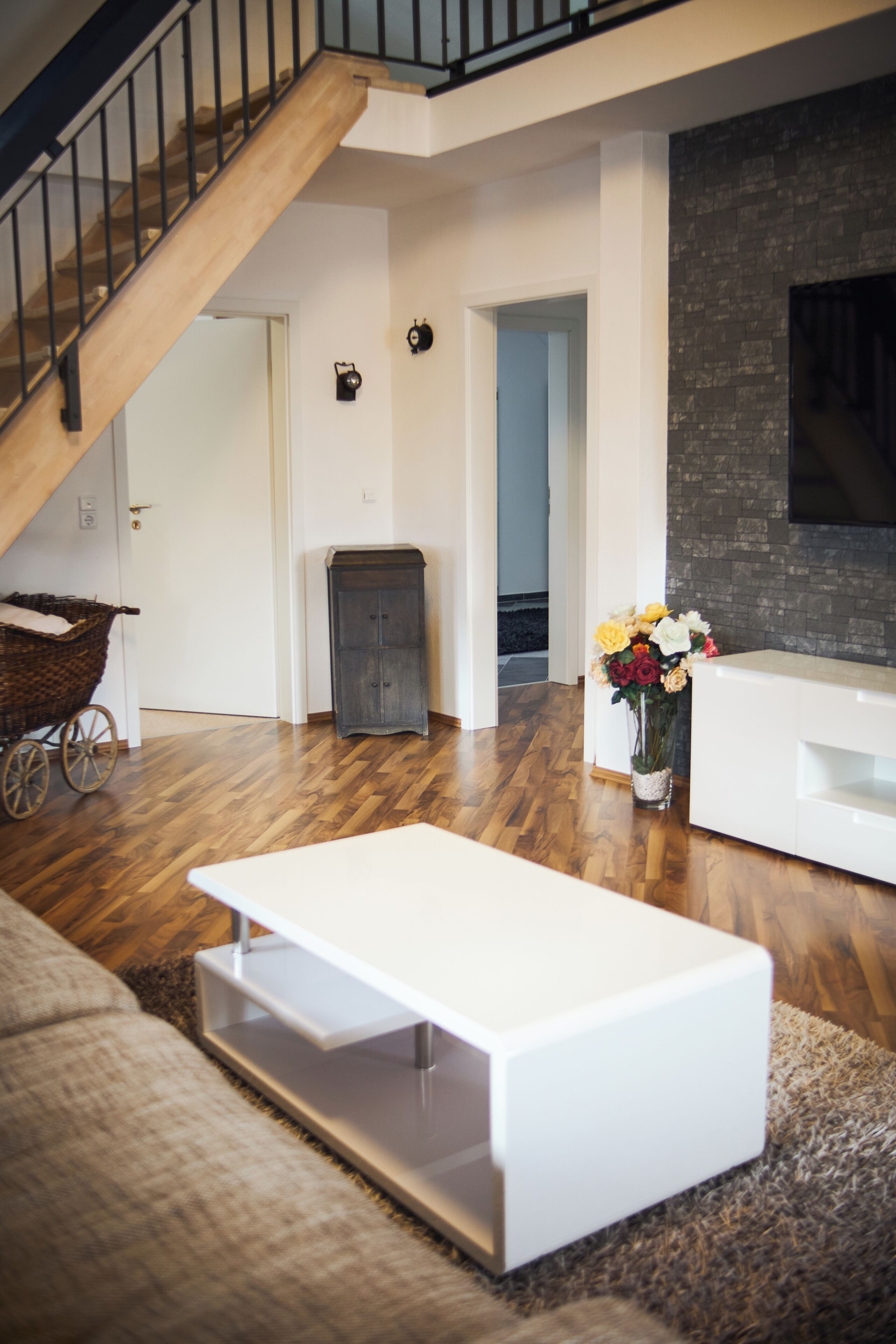 A modern living room with a wooden staircase, a white sofa, a white coffee table, a grey cabinet, and a vase of flowers.