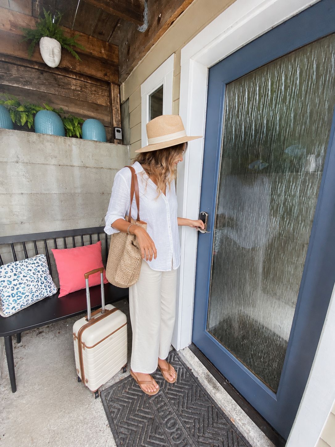 Una persona de pie frente a una puerta, con una maleta y un bolso, un banco con cojines y una pared decorada.