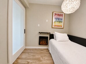 A bedroom with a fireplace, a bed with white bedding, a wooden floor, and a wall-mounted light fixture.