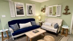Ein Wohnzimmer mit einem blauen Sofa, einem weißen Sessel, einem Holztisch und zwei gewobenen Hocker. An den Wänden befinden sich gerahmte Bilder und ein kleiner Tisch mit einer Lampe.
