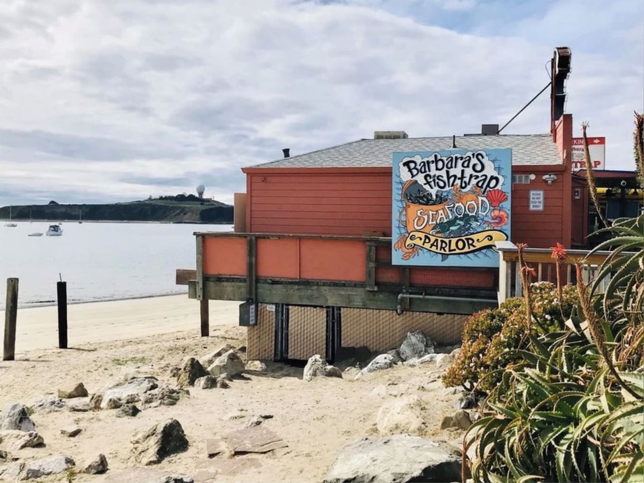 Een restaurant aan het strand met een bord waarop zeevruchten staan aangeboden en uitzicht op de oceaan.