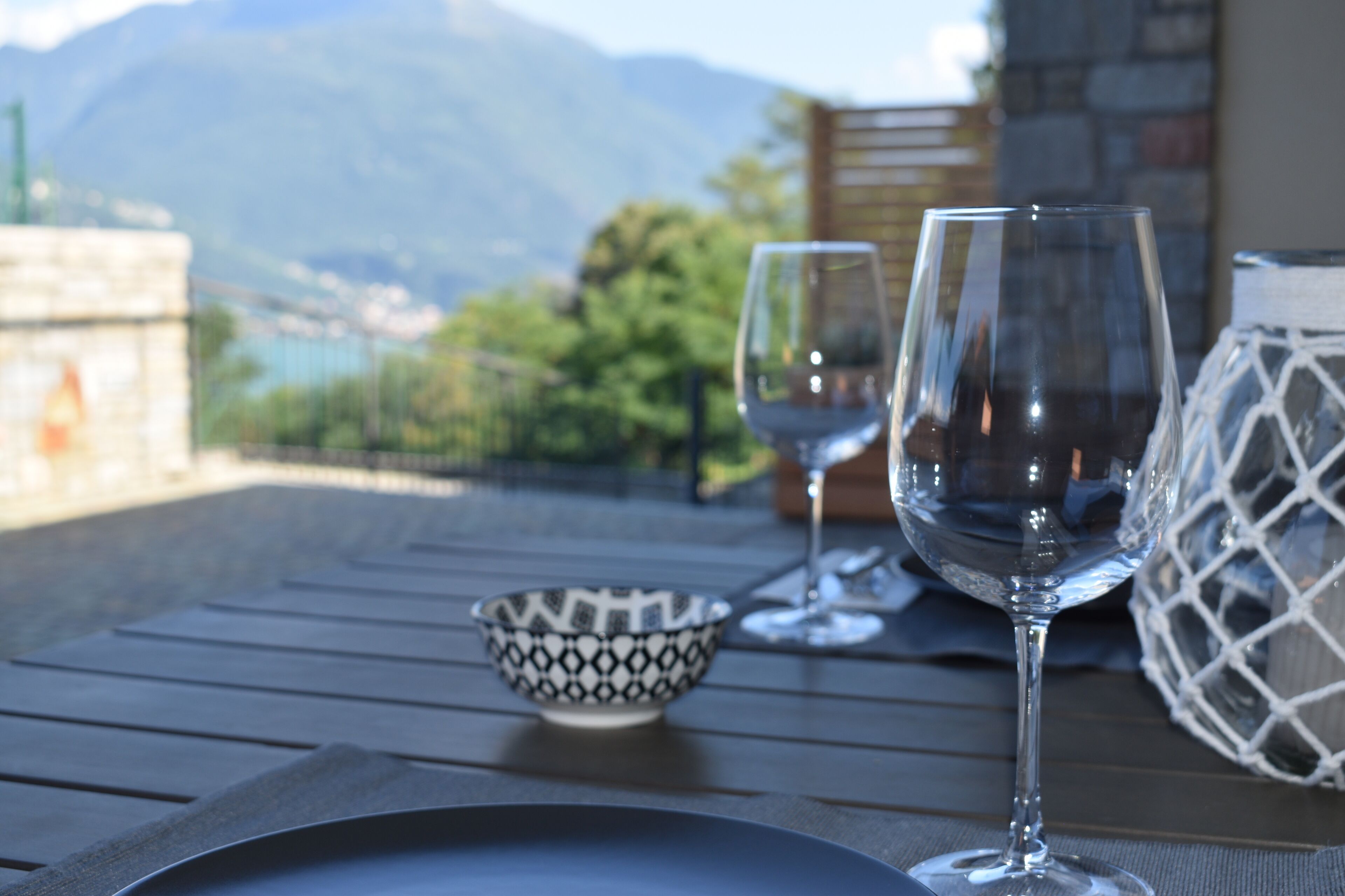 A dining table with two wine glasses and a patterned bowl, set against a backdrop of mountains and a stone wall.