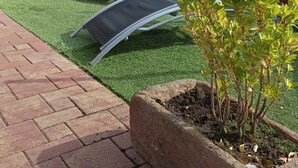 Uma área de jardim com grama sintética, cadeiras de descanso e um vaso de planta.