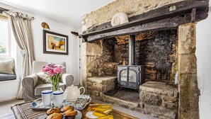 A cozy living room with a stone fireplace, a wicker sofa, a dining table with a tray of muffins, and a framed picture on the wall. - Willow Cottage, Chilmark, Wiltshire (Chilmark)