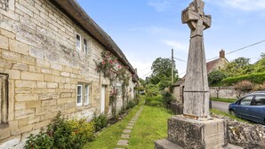 A stone building with a thatched roof, a narrow stone pathway, and a wooden cross monument. - Willow Cottage, Chilmark, Wiltshire (Chilmark)