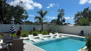 A swimming pool with lounge chairs and a table, surrounded by a white fence and greenery. - Bradenton Gem - Beaches, Downtown, IMG Academy, New Pool! (Bradenton)