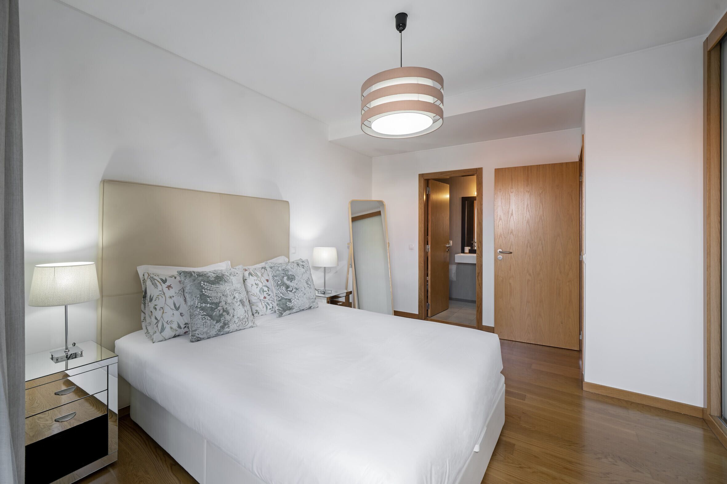 A modern bedroom with a large bed, bedside tables, a mirror, and a wooden door leading to a bathroom.